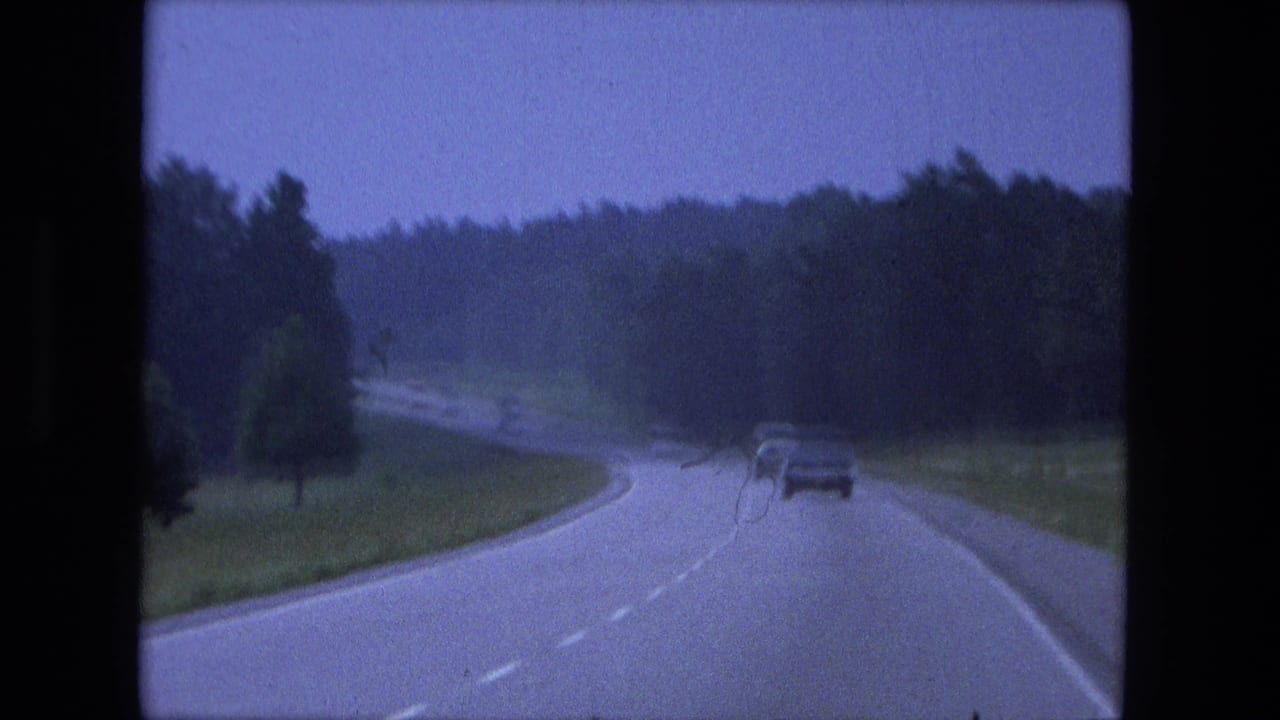 Home-movie footage shows a moving-vehicle perspective on a wooded Indiana highway, with one car ahead near the curve.