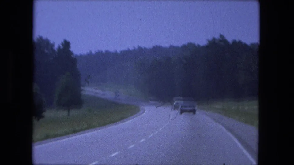 Home-movie footage shows a moving-vehicle perspective on a wooded Indiana highway, with one car ahead near the curve.
