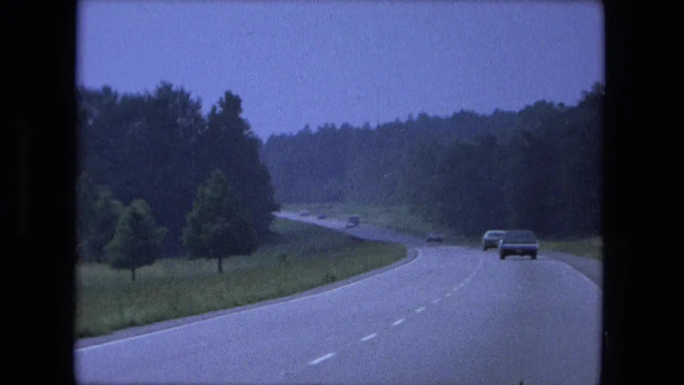The 8mm frame shows a 1970s road-travel view near Fort Wayne, with a car ahead on a forest-lined divided highway.