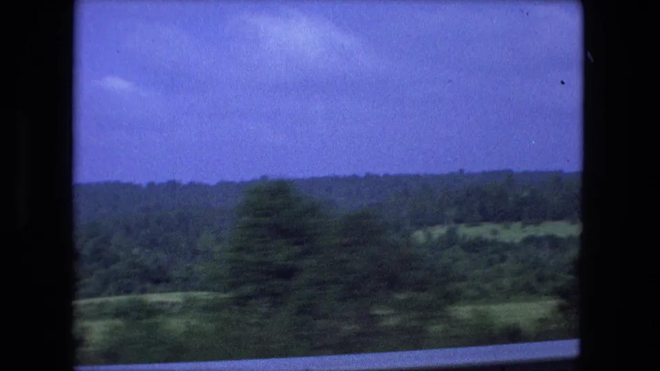 A moving-vehicle view captures blurred wooded landscape and rolling terrain, consistent with the same 1974 home-movie road sequence.