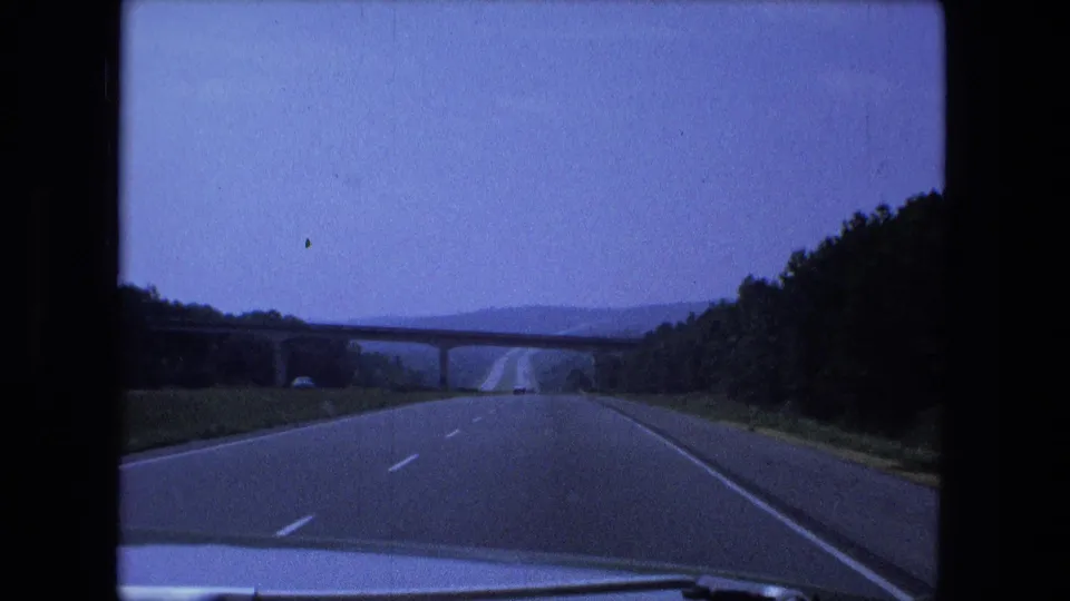 1974 Fort Wayne area highway view with a concrete overpass ahead, tree-lined shoulders, and hazy blue sky.