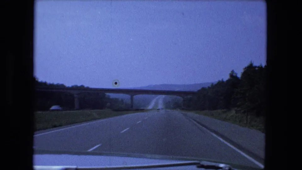 Moving-vehicle 8mm view of an Indiana highway, overpass, trees, and distant low hills in 1974.