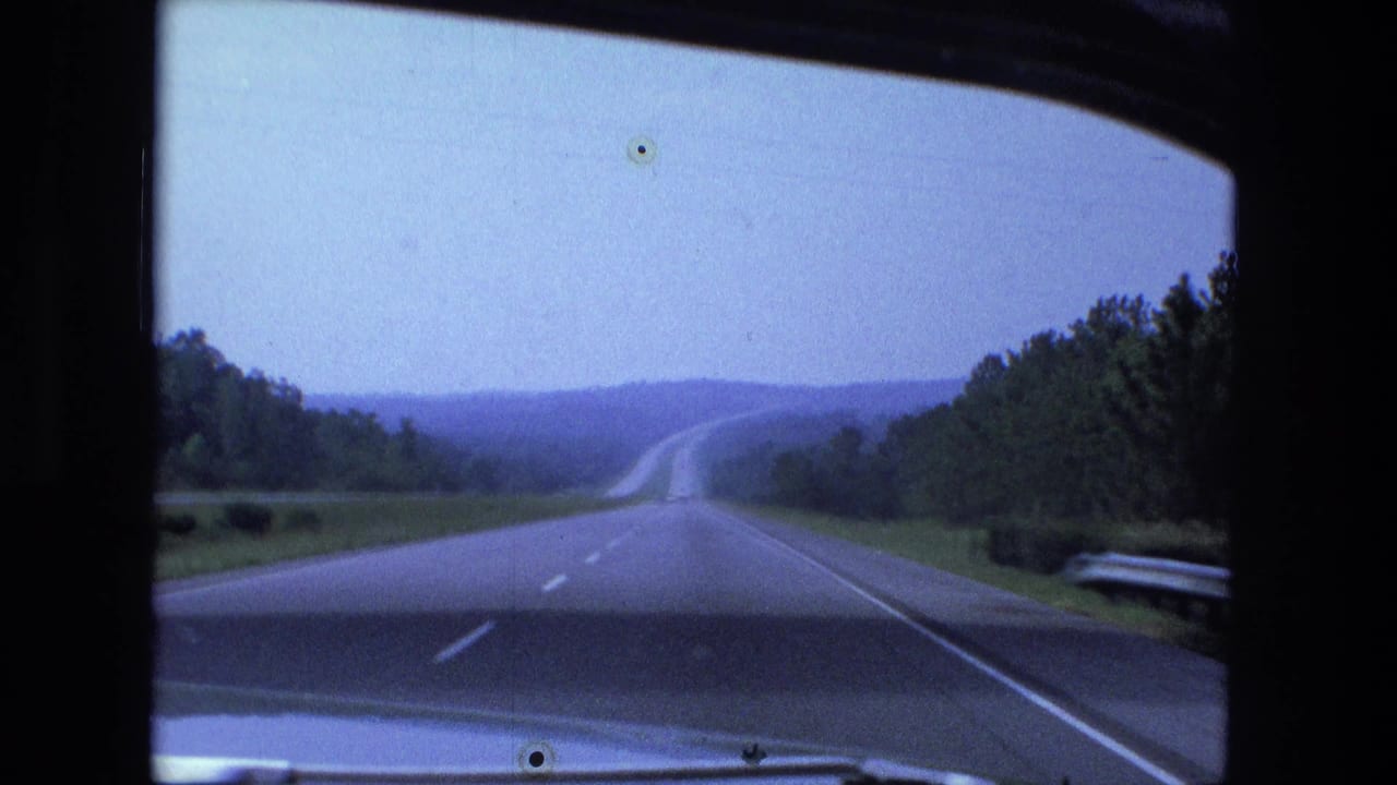 Straight highway seen through a car windshield with trees, hills, lane markings, and part of the hood visible.