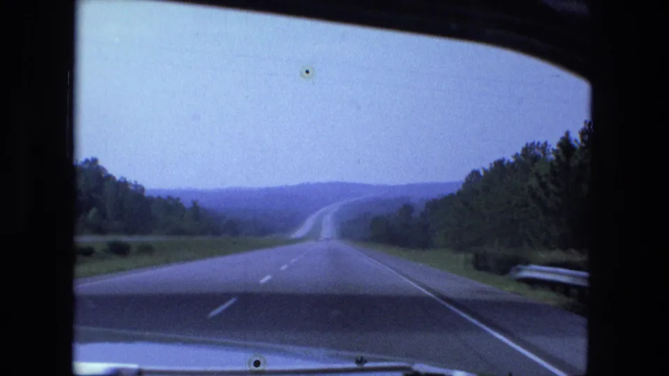 1974 Fort Wayne-area home-movie frame showing a long highway through green roadside landscape from inside a moving car.