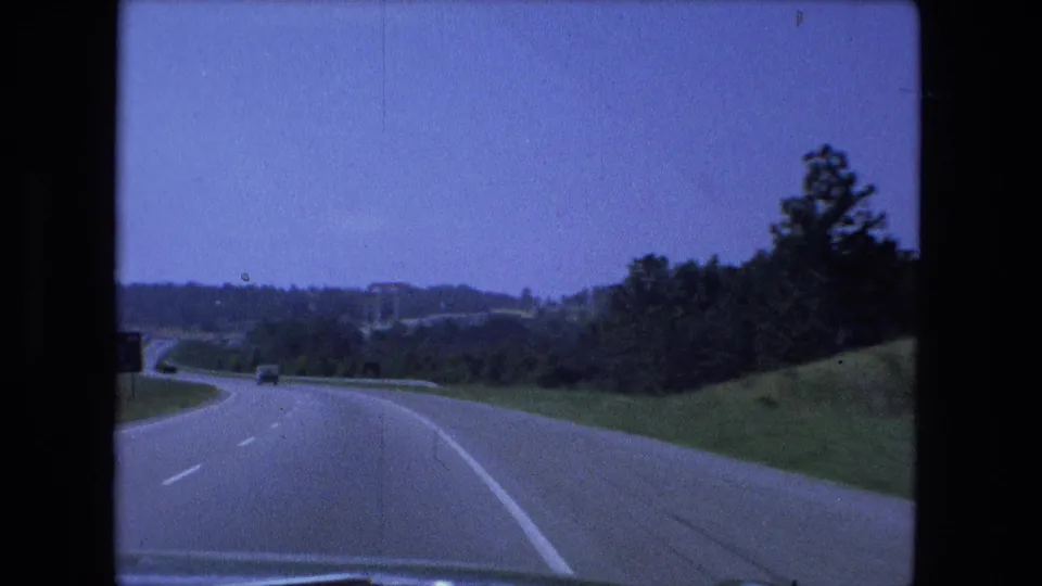 Fort Wayne-area 1974 home-movie frame of a highway curve with sparse traffic and green roadside terrain.