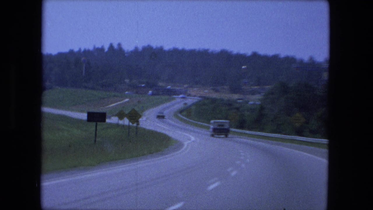 Winding divided highway with cars, roadside signs, and wooded slopes beyond the road.