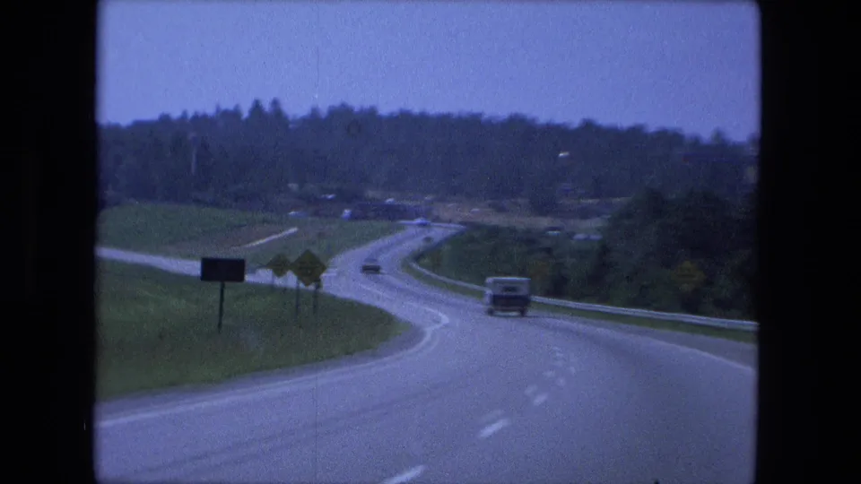 1974 Fort Wayne-area road-trip footage showing a winding divided highway with cars, signs, grassy shoulders, and trees in the distance.