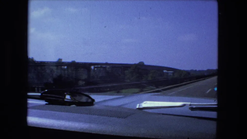Dashboard view from 1974 road-trip footage as the car approaches or crosses a bridge, with guardrails and road surface visible ahead.