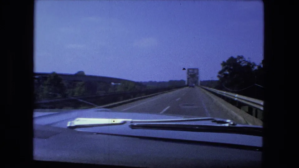 1974 Fort Wayne-area dashboard shot showing the car traveling on a bridge or raised road behind a vehicle, with guardrails and sky visible.