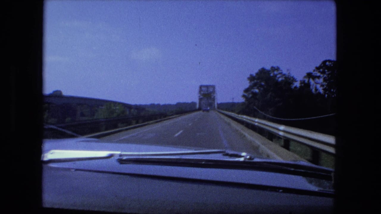 Car approaches a distant steel truss bridge on a two-lane road, with dashboard and wipers visible.