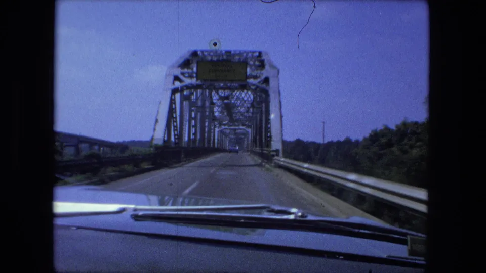 Inside-car view approaching the portal of a steel truss bridge in Fort Wayne, with road surface, guardrails, trees, and dashboard edge visible.
