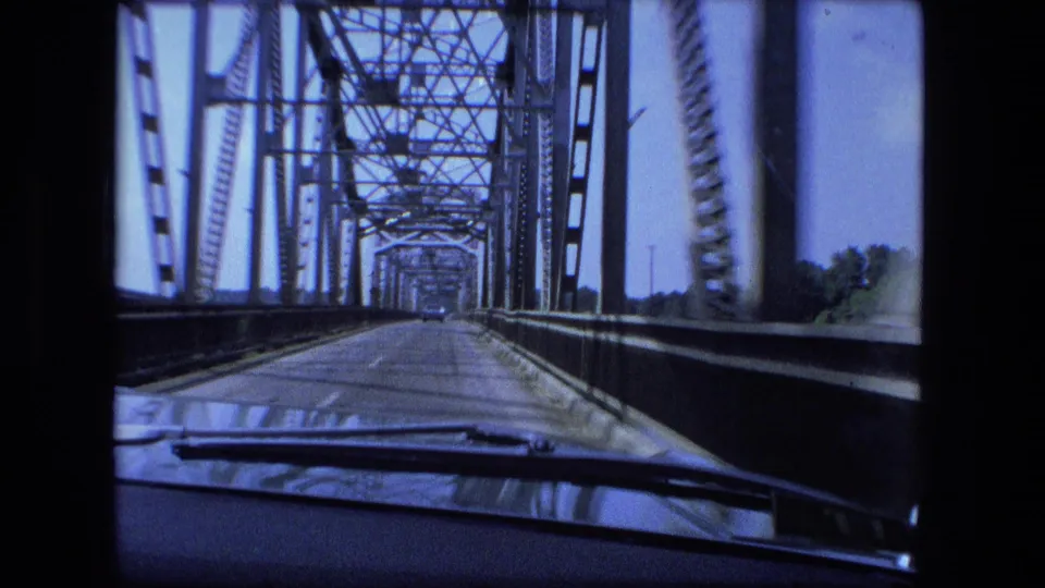 Point-of-view 8mm frame from inside a car crossing a steel truss bridge, showing repeating beams, side railings, road deck, and windshield wipers.