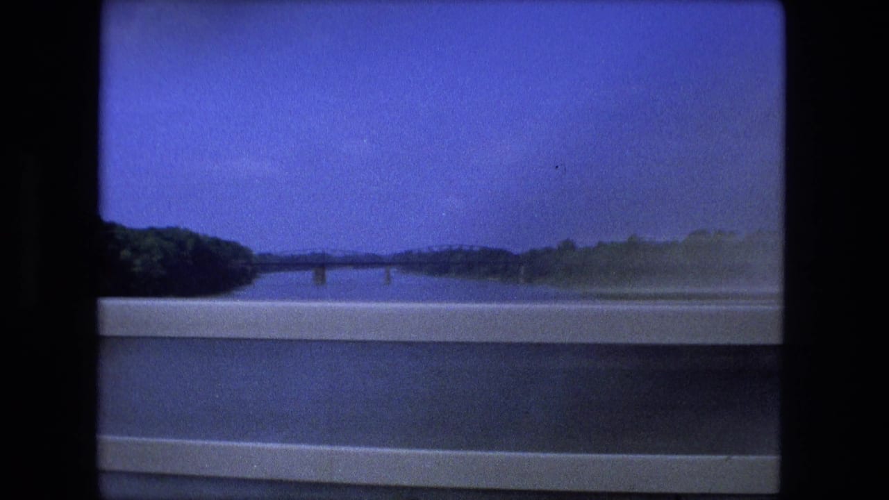 1974 Fort Wayne frame looking downriver from a bridge, with guardrails in the foreground and wooded banks under a deep blue sky.