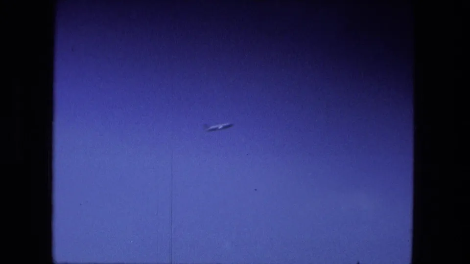 1974 Fort Wayne frame showing a small aircraft-like shape against blue sky, with film grain and the bridge crossing no longer prominent.