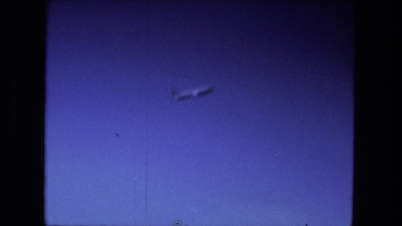 Small airplane crossing a deep blue sky in a dark 8mm film frame.
