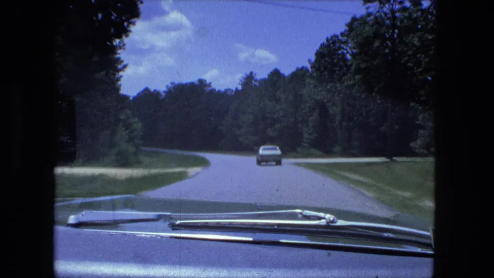 A 1974 home-movie road view shows a car ahead on a curving, tree-lined rural road with the filming car hood visible.
