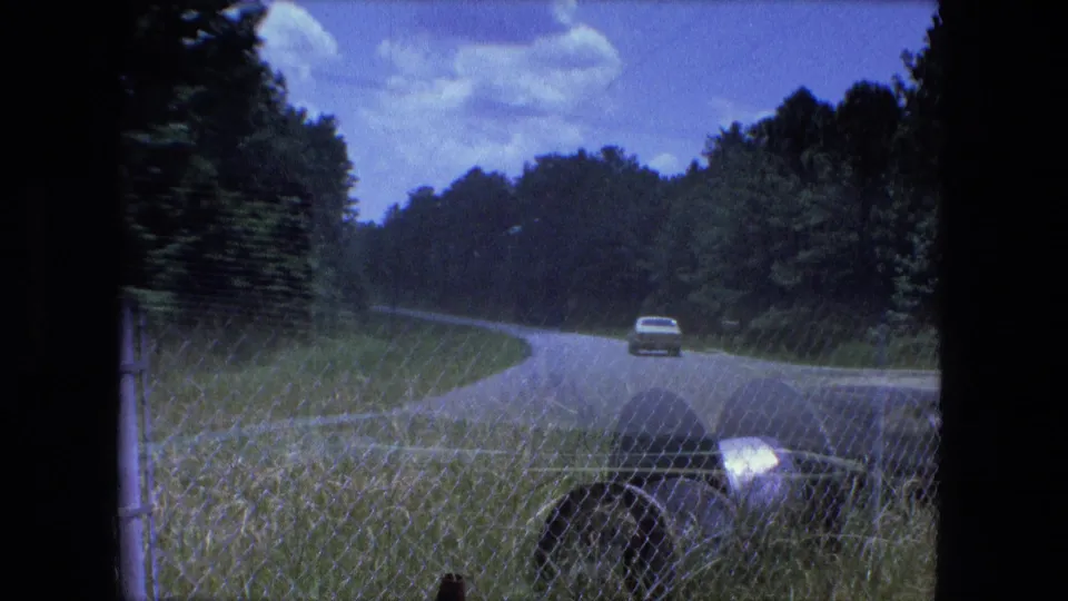 A distant man walks in a grassy area behind or near a chain-link fence, with large wire spools visible in the foreground of the Indiana reel.