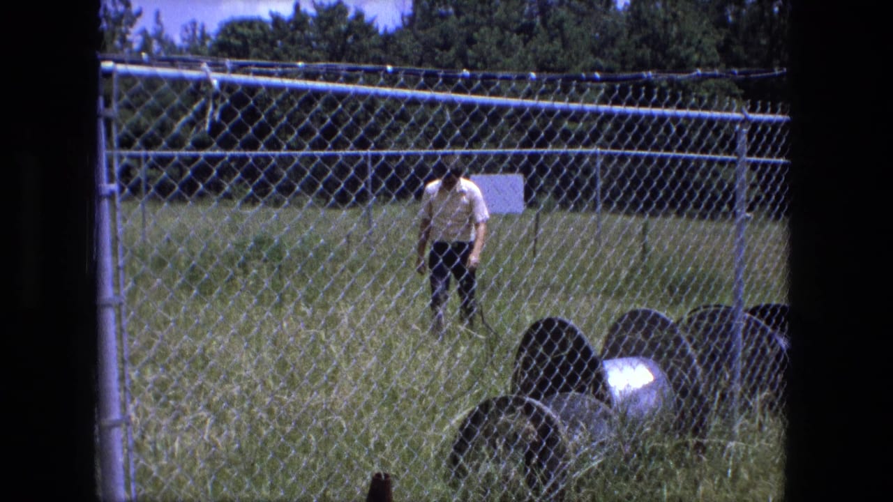 Man in a light shirt walks through tall grass behind a chain-link fence.