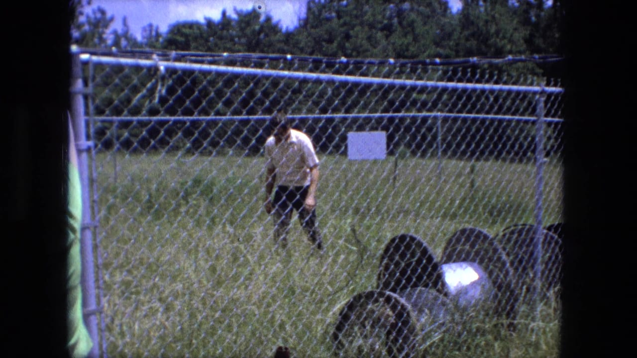 Man walks along the grassy fenced area with trees behind him.