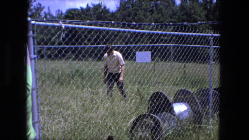 The man continues across the Fort Wayne grassy enclosure in 1974, framed through chain-link fencing with dark round objects in the foreground.