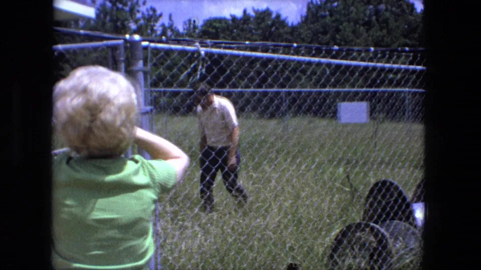 A woman in a green shirt watches from near the camera while the man approaches through the fenced Fort Wayne yard in 1974.