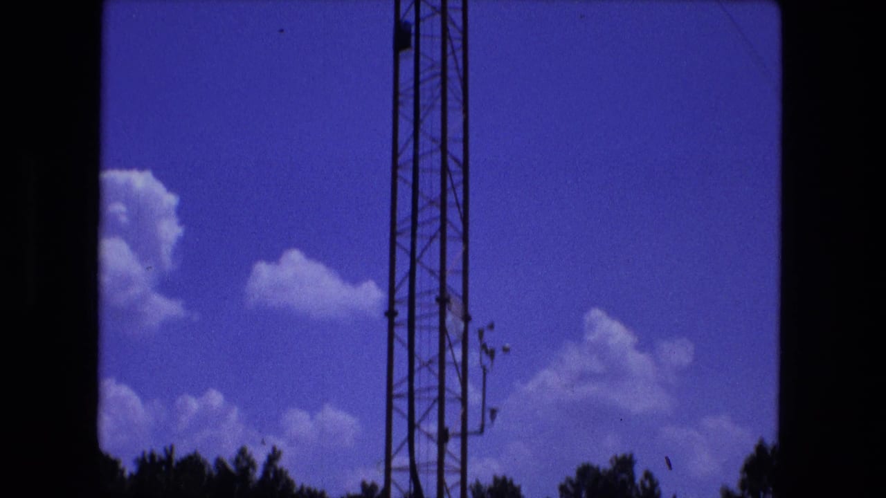 Lattice antenna tower rising against blue sky with white clouds and dark treetop edge.
