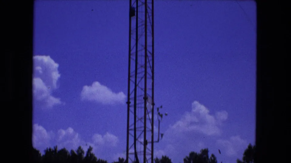 1974 Fort Wayne 8mm frame showing a tall lattice antenna tower, guy wires, blue sky, clouds, and trees near the bottom edge.