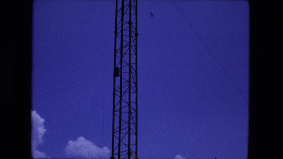 1974 Fort Wayne home-movie frame with the tower shifted to the left of center, showing wires, blue sky, and small clouds.