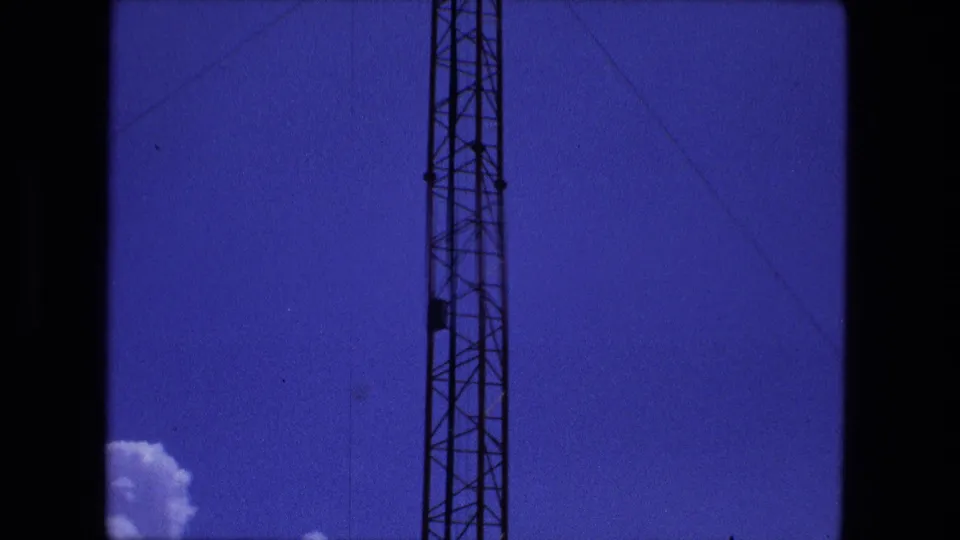 1974 Fort Wayne 8mm frame showing a vertical antenna tower, thin guy wires, blue sky, and a small cloud at the bottom corner.