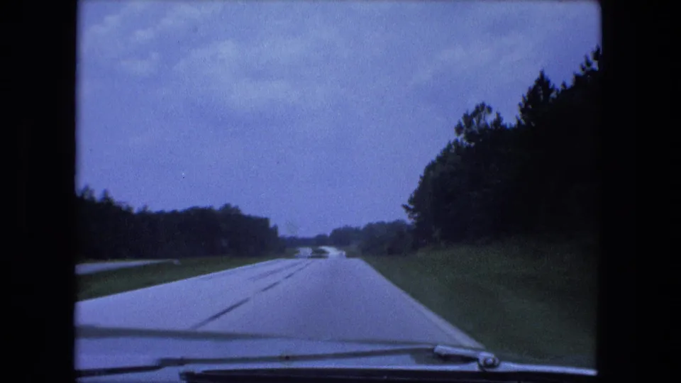 1974 Fort Wayne-area home-movie view from a moving car on a lightly traveled divided highway with trees, grass, and pale sky.