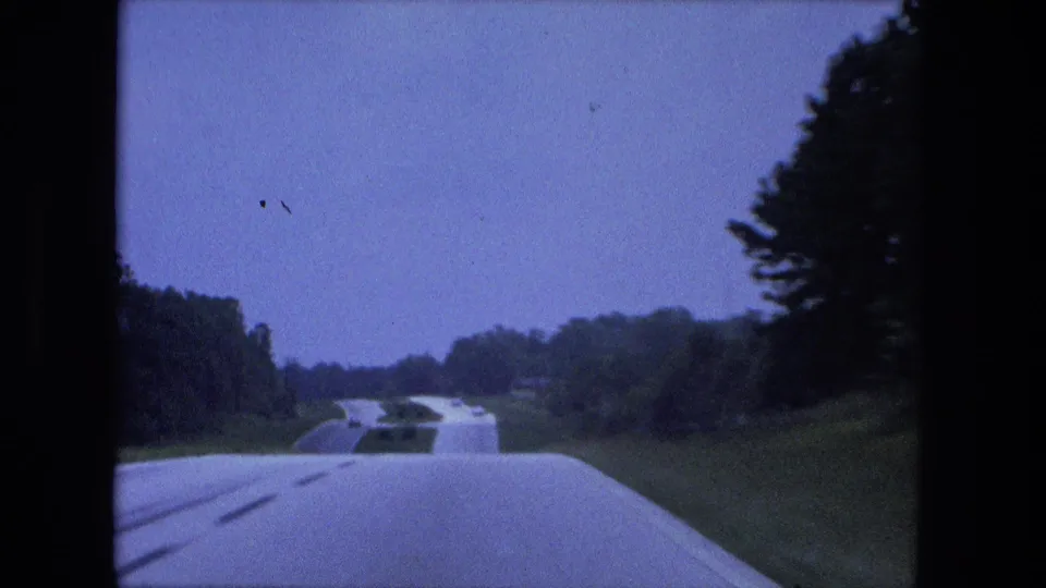 Home-movie frame of a 1974 Fort Wayne-area interstate seen from inside a car, with a curving road, grassy median, and sparse traffic.
