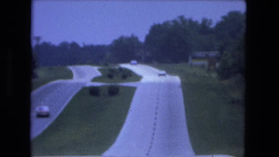Fort Wayne-area 1974 8mm frame showing a lightly traveled divided highway, green median, roadside trees, and distant cars.