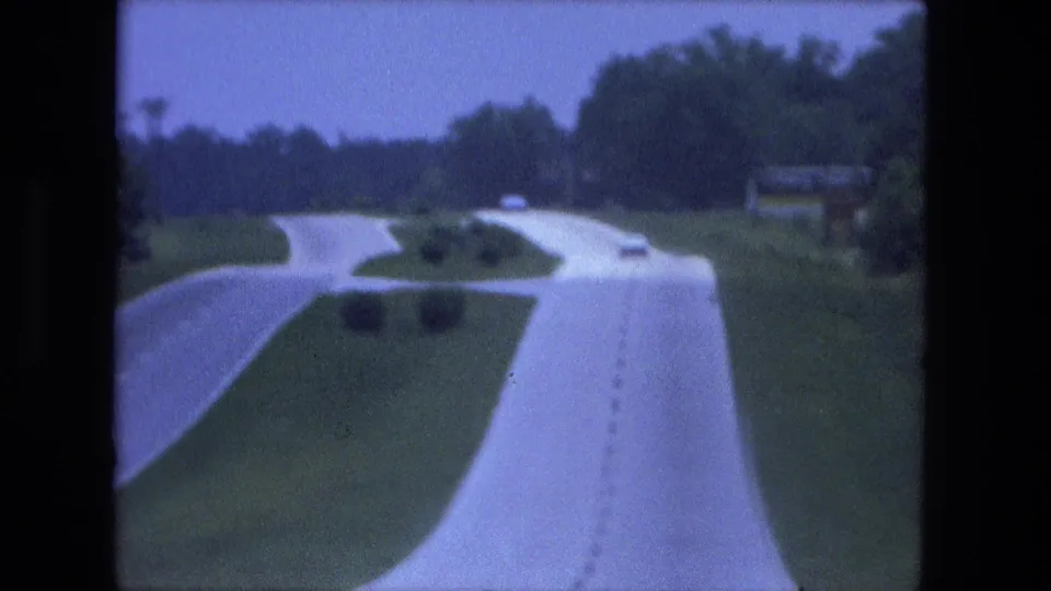 1974 8mm home-movie view from a moving car on a curving divided road near Fort Wayne, Indiana, with grass medians and distant vehicles.
