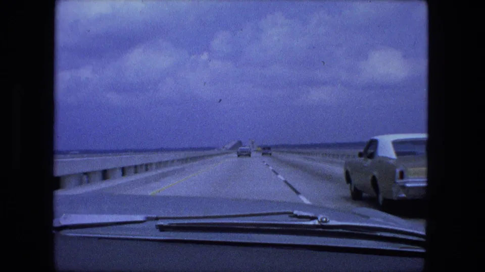 1974 8mm driving view from inside a car on a long multi-lane bridge or causeway, with traffic ahead and an older car in the adjacent lane.