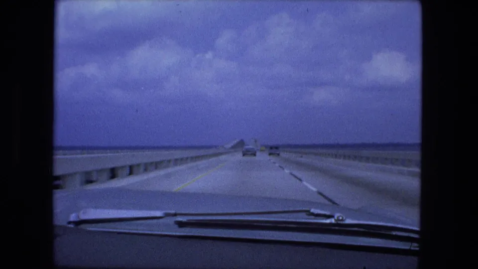 Windshield-level 1974 home-movie frame showing a long bridge or causeway roadway, concrete rails, lane markings, and traffic in the distance.
