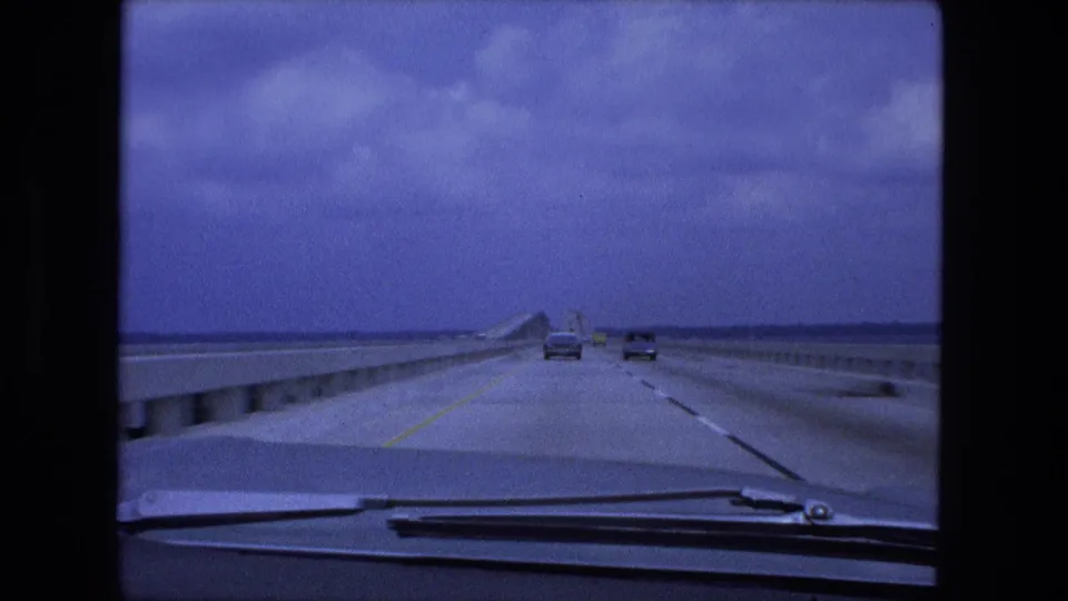 A 1974 8mm frame from a moving car shows a bridge-like interstate stretch, concrete barriers, two vehicles ahead, and a blue-tinted cloudy sky.