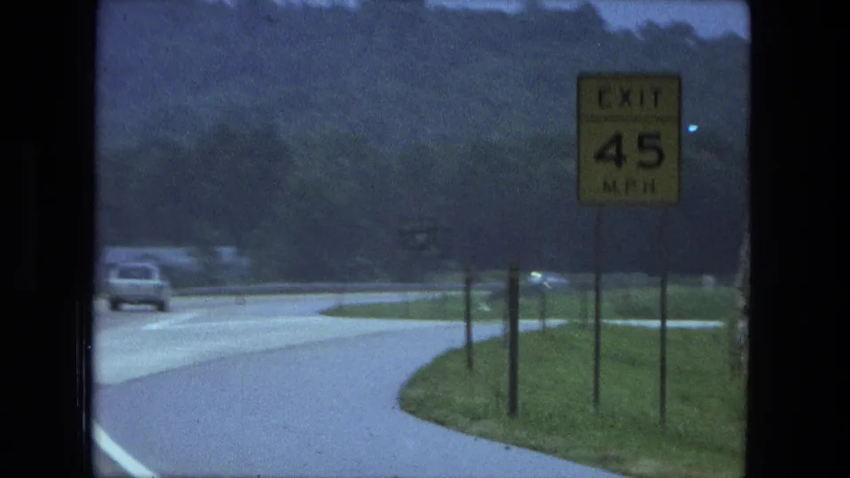 1974 Fort Wayne area 8mm frame showing a curved rural highway, yellow exit speed sign, grass verge, trees, and distant cars.