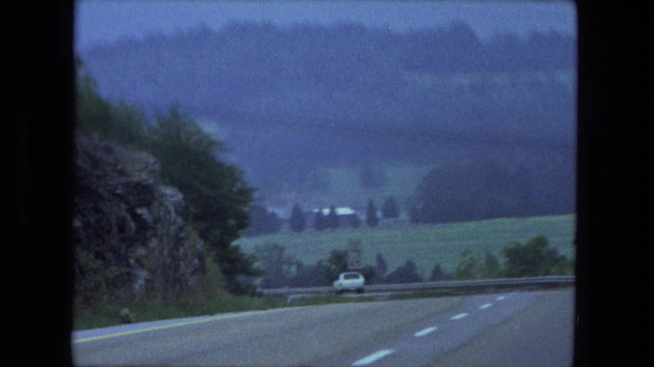 Two-lane highway running downhill past a rocky or wooded roadside toward fields and trees.