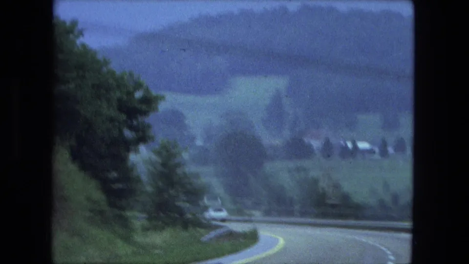 1974 Fort Wayne area frame with a sweeping highway curve, guardrail, trees, and hazy green fields in the distance.