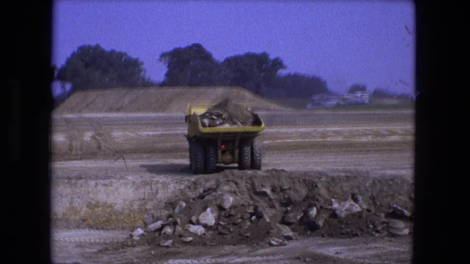 In 1974 Fort Wayne, Indiana footage, a yellow dumper truck moves across a graded construction site toward a pre-dug hole.