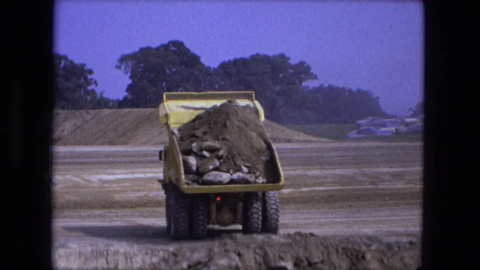The yellow dumper truck is seen from behind at the Fort Wayne construction site, positioned near the excavation with its load still raised inside the bed.