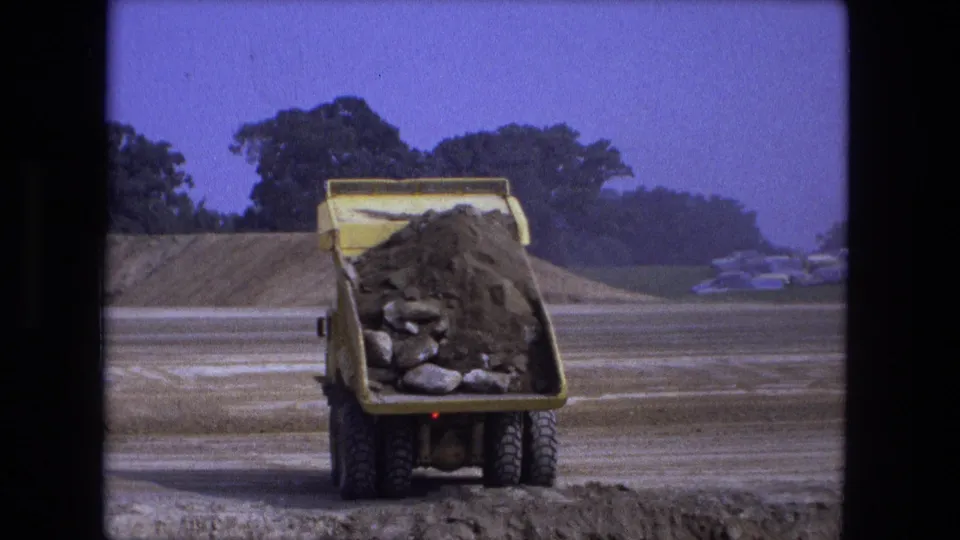 At the 1974 Indiana worksite, the dumper truck begins tipping soil and rocks from its bed into the pre-dug hole.