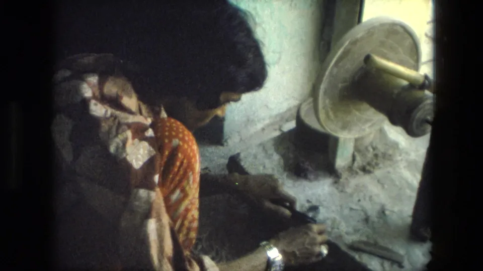 In 1974 India, a woman in a patterned sari appears to work at a stone craft station beside a grinding wheel.