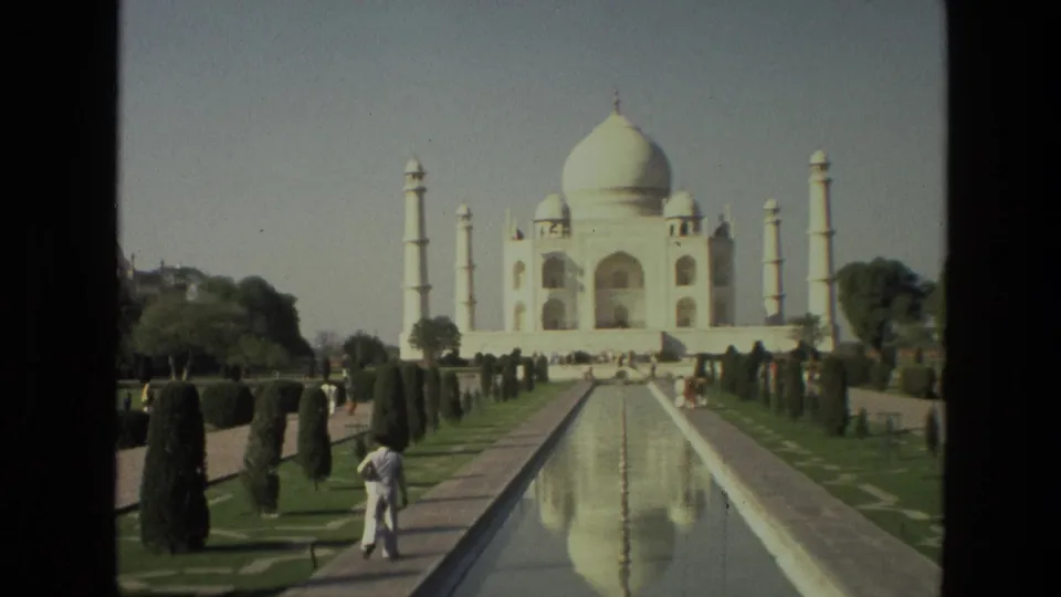 1974 India home-movie frame of the Taj Mahal, its dome and minarets reflected in the central water channel as a visitor walks nearby.