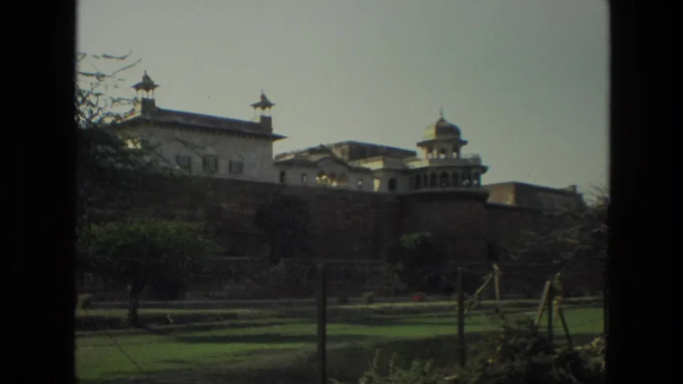 1974 India travel frame showing a large historic fort exterior with stone walls, domed structures, lawns, and trees.