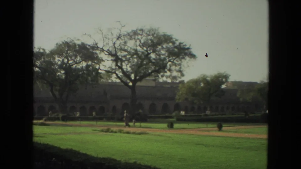1974 India 8mm frame showing a long arched building, manicured lawns, mature trees, and distant visitors across the grounds.