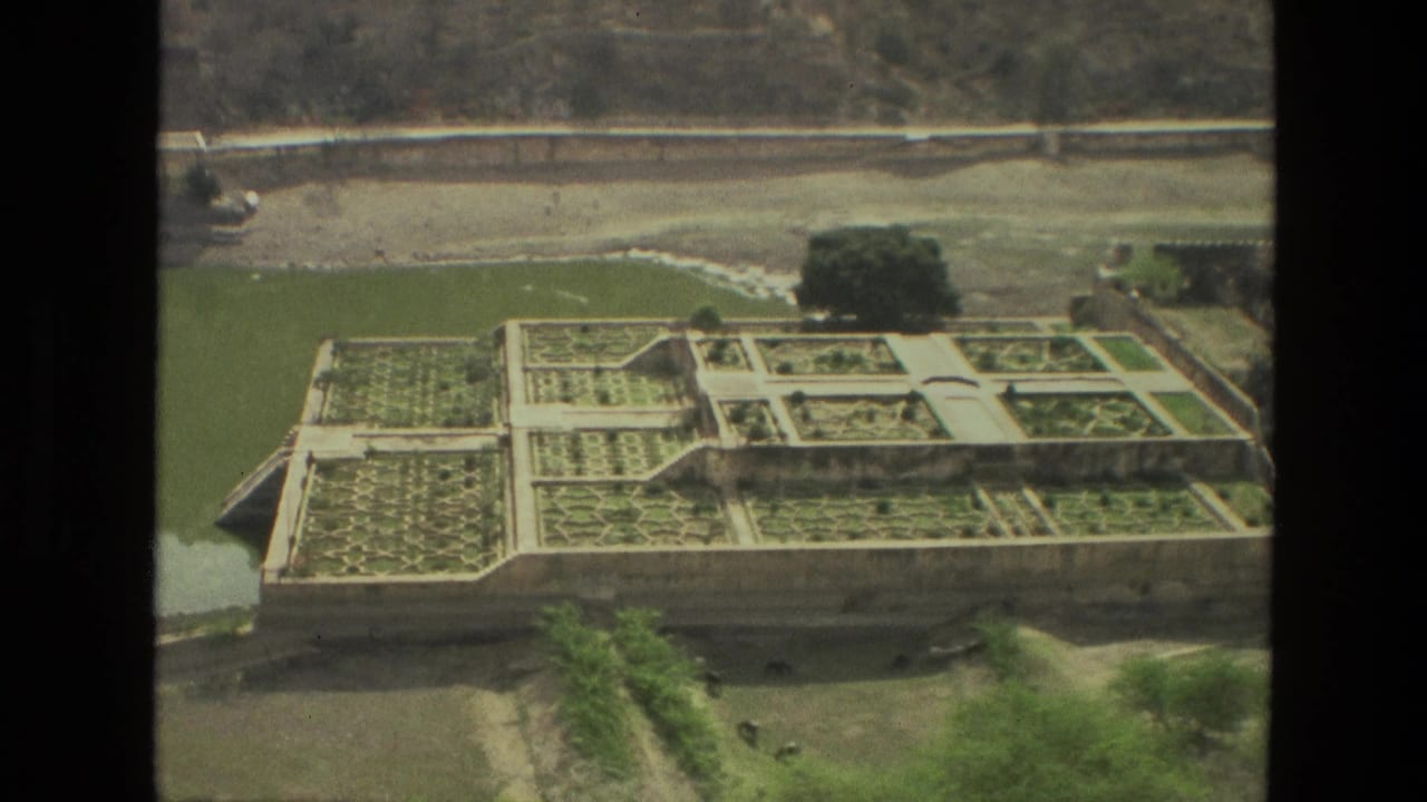Elevated view of a formal terraced garden beside a green pond and road.