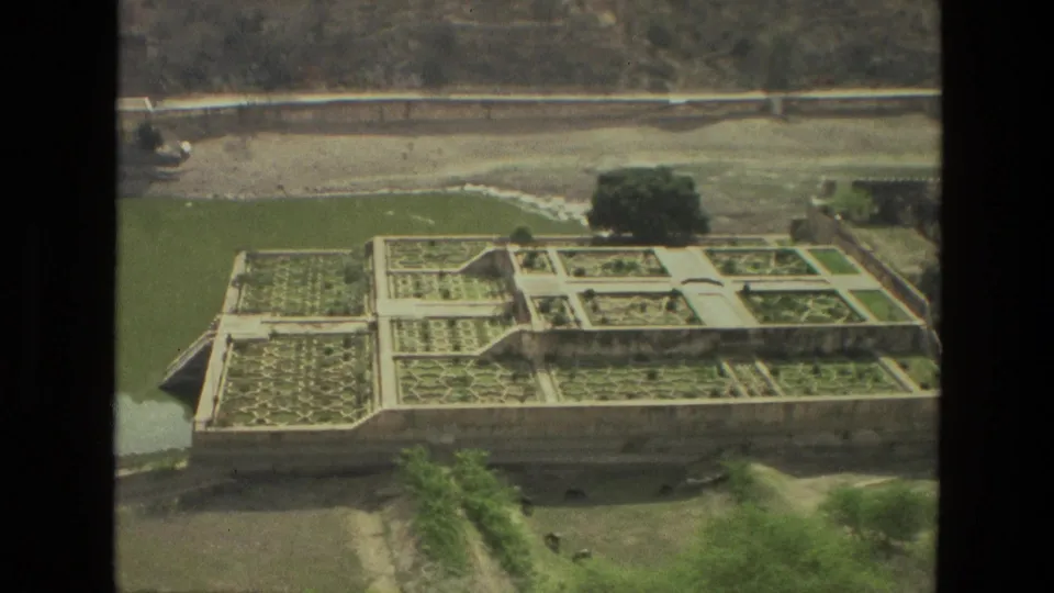 1974 India 8mm frame showing a formal terraced garden, pale paths, masonry edges, green water, trees, and a distant road.