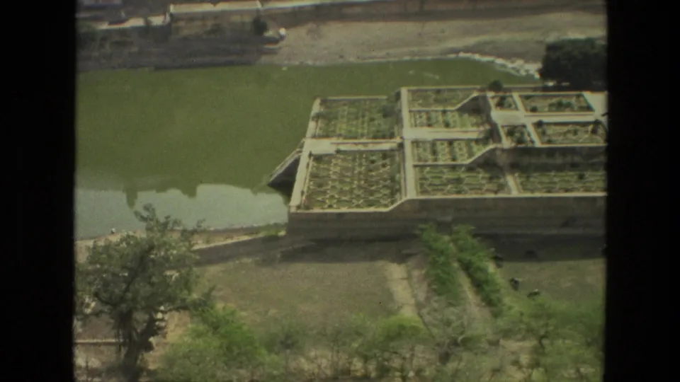 1974 India 8mm frame drifting along the garden edge, with green pond water, terraced beds, trees, and exposed ground below.