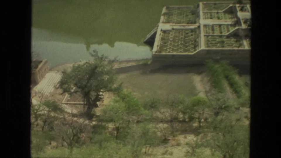 1974 India home-movie frame showing the corner of a terraced garden, pond water, trees, paths, and dry open ground from above.